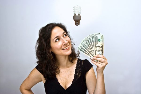 Woman waving money and looking up. Having an environmentally friendly idea with an energy saving flourescent light bulb floating above her head.